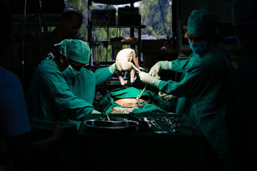 Medical team performing surgery in a dimly lit operating room, with a newborn being held above the surgical area.
