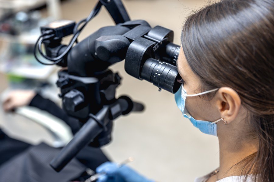 Female dentist wearing a surgical mask examines a patient’s teeth through a dental microscope.